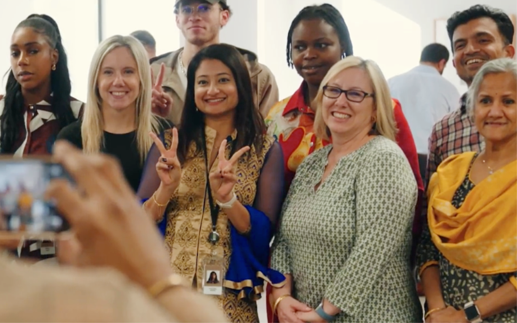 Group of colleagues posing for a photo at an office celebration; the person in the center wears a gold‑and‑blue traditional outfit and holds up two peace signs, surrounded by others in colorful patterned attire, with a hand holding a smartphone visible in the foreground.